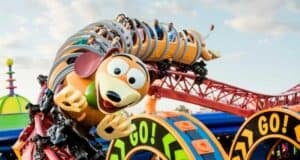 A colorful roller coaster at Disney World, shaped like a springy dog with a cartoonish face, is full of excited riders. The track features bright, playful designs, including loops marked "GO!" against a sunny sky.