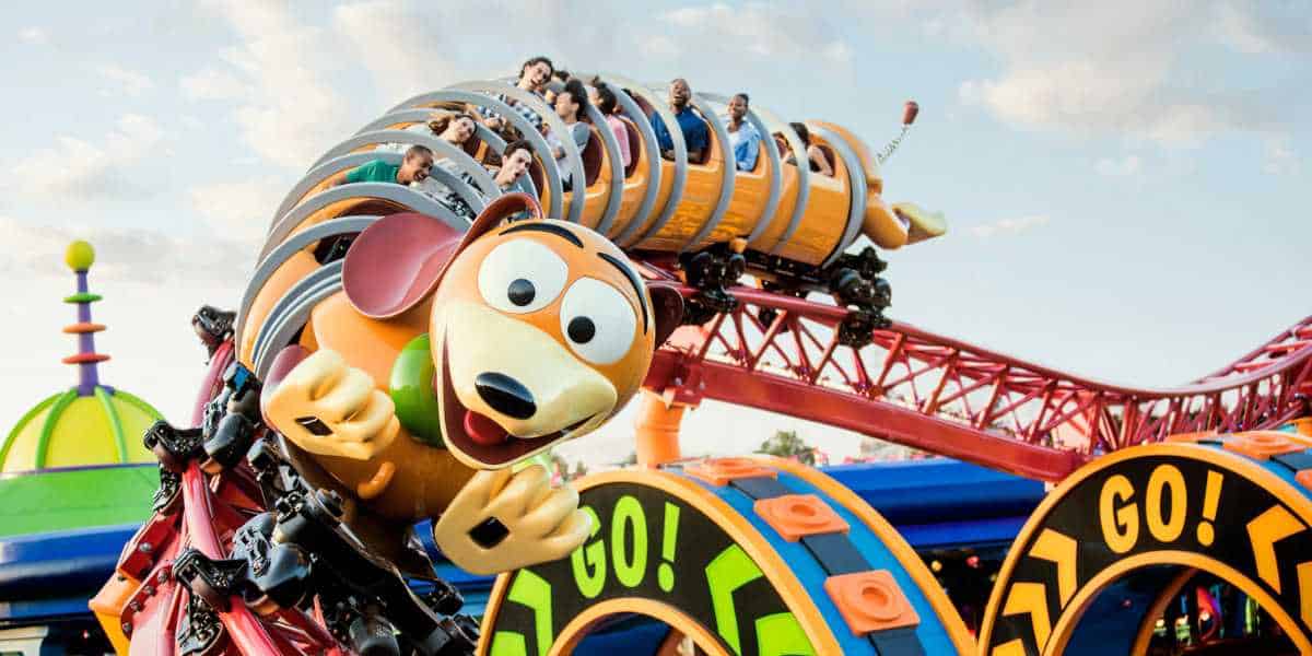 A colorful roller coaster at Disney World, shaped like a springy dog with a cartoonish face, is full of excited riders. The track features bright, playful designs, including loops marked "GO!" against a sunny sky.