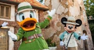 Two Disney characters dressed as explorers stand cheerfully outside a rustic building. The character on the left wears a green safari outfit, and the one on the right wears a beige explorer outfit with a hat. Trees and a wooden structure are in the background.