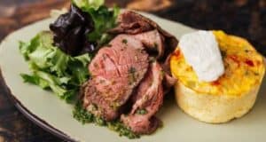 A plate with slices of roast beef in herb sauce, a serving of leafy greens, and a small frittata topped with a dollop of cream.