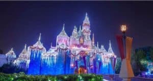 A brightly illuminated castle at night, adorned with twinkling lights in various colors. The sky is clear and dark, enhancing the vibrant display. Foreground features a stone pathway and lush greenery.