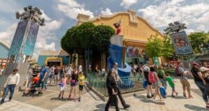 A bustling scene at Walt Disney Studios with a large crowd of people, some pushing strollers. The main building features Disney-themed decorations. Bright, sunny weather with a partly cloudy sky and lush greenery surrounds the area.