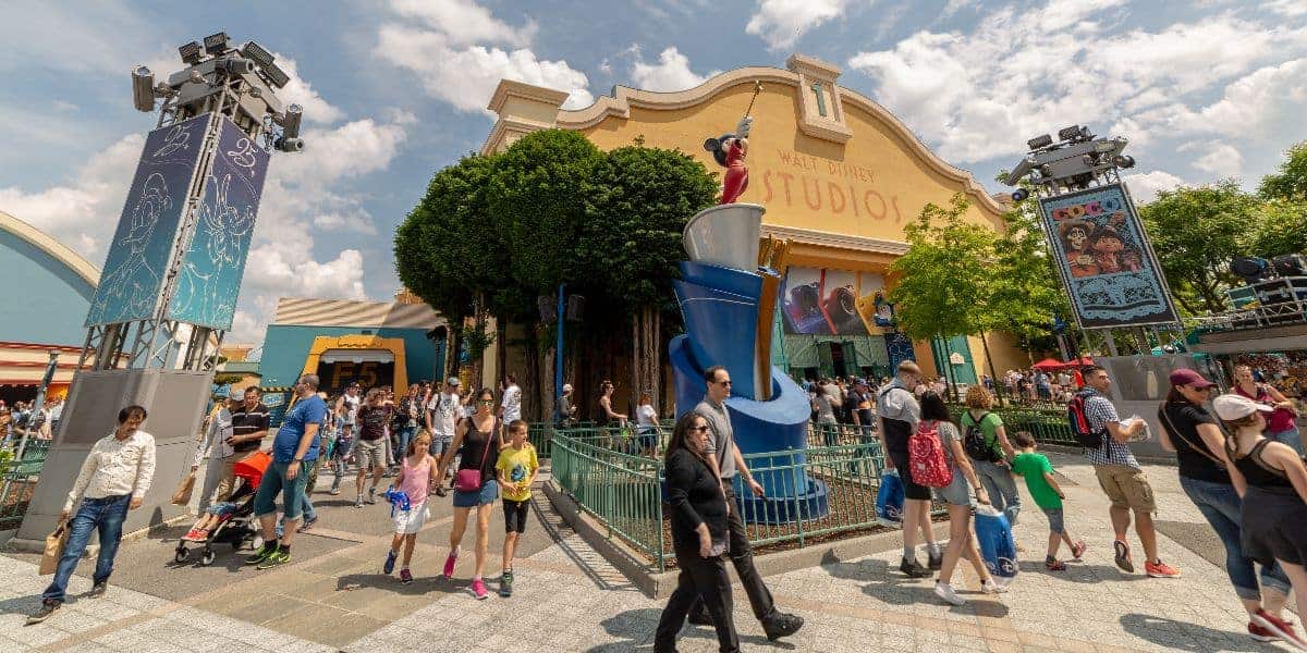 A bustling scene at Walt Disney Studios with a large crowd of people, some pushing strollers. The main building features Disney-themed decorations. Bright, sunny weather with a partly cloudy sky and lush greenery surrounds the area.