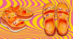 Bright orange platform sandals adorned with colorful charms, including rainbows and flowers, are displayed against a vibrant pink and yellow wavy background.