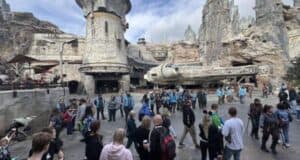 A bustling theme park scene with people gathered around a large, detailed spacecraft and rugged, rocky structures. The sky is partly cloudy, enhancing the adventurous atmosphere of the location.