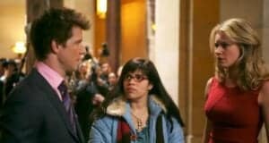 A man in a suit and pink shirt talks to a woman with glasses and braces, wearing a blue coat with a colorful scarf. Another woman in a red dress looks on. They are indoors with people and cameras in the background.