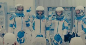 Four astronauts in blue and white space suits wave and pose for a photo in a control room. Equipment and screens are visible in the background, and people in white attire and caps are taking pictures.
