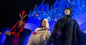 Three people dressed as iconic villains stand in front of a castle lit in blue. On the left, a figure in red with a tall hat; in the center, wearing a fur coat; and on the right, in black with horned headgear.