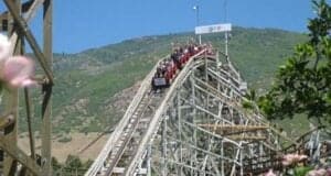 A roller coaster car filled with passengers descends a wooden track under a clear blue sky. The track is surrounded by greenery and a mountain landscape in the background. Pink flowers are visible in the foreground.