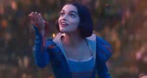 A person wearing a blue and red medieval-style dress is gazing upwards, smiling, with one hand extended as if reaching for something. The background is a softly blurred natural scene with warm, glowing lights.