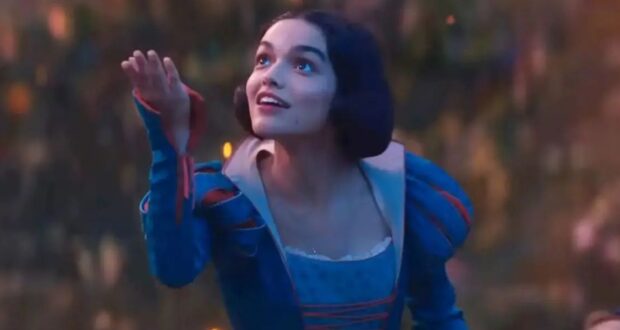 A person wearing a blue and red medieval-style dress is gazing upwards, smiling, with one hand extended as if reaching for something. The background is a softly blurred natural scene with warm, glowing lights.