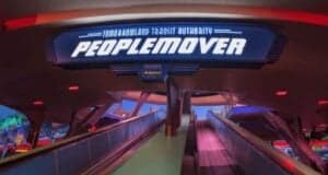 Sign for the Tomorrowland Transit Authority PeopleMover at an amusement park, above an ascending walkway. The area is illuminated with colorful lights, creating a futuristic atmosphere.
