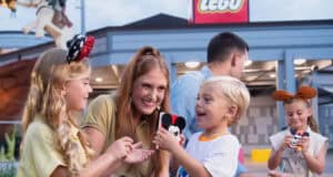 A smiling woman and two children hold Mickey Mouse-themed LEGO creations outside a LEGO store at Disney Springs. In the background, a man and girl wearing mouse ears enjoy more Disney Springs activities, building with colorful LEGO bricks.