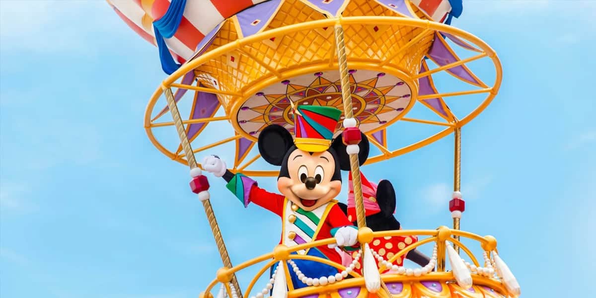 Mickey Mouse, dressed in a colorful festive outfit, stands on a vibrant, elaborate parade float designed like a hot air balloon basket, waving against a clear blue sky.