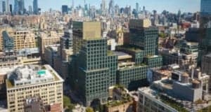 Aerial view of a cityscape featuring modern high-rise buildings with green terraces. The background displays a sprawling urban skyline with numerous skyscrapers under a clear blue sky.