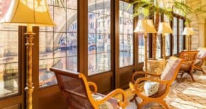 Sunny indoor seating area with elegant wooden chairs, patterned cushions, tall lamps, potted palm plants, and large windows offering a view of a picturesque canal and arched stone bridge outside.