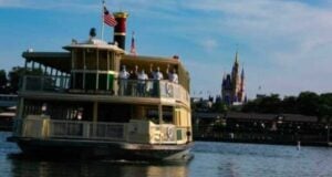 A ferryboat named Admiral Joe Fowler crosses a lake with several people on board; American flags are flying, and a castle is visible in the background under a blue sky.