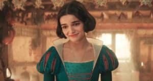 A woman with short dark hair smiles softly. She is wearing a medieval-style dress with puffed sleeves and intricate teal accents. The background features a warmly lit room with hanging decorations.
