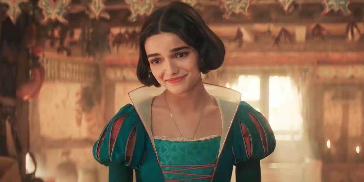 A woman with short dark hair smiles softly. She is wearing a medieval-style dress with puffed sleeves and intricate teal accents. The background features a warmly lit room with hanging decorations.