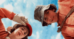 Two teens in orange uniforms and hats look down toward the camera against a blue sky, appearing curious or puzzled. One wears gloves and adjusts their red cap, while the other wears a bucket hat.