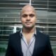 Professional headshot of Devraj in a formal suit, standing in an office environment.