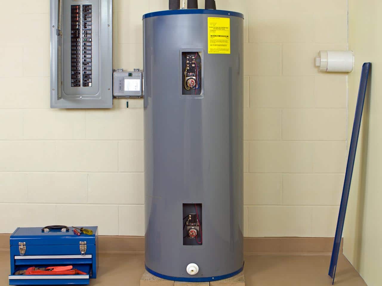 A large gray water heater stands in a utility room, ready for winter maintenance, with a closed electrical panel on the wall and a blue toolbox on the floor.