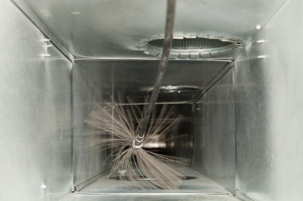 A brush and hose inside a metal air duct, viewed head-on, emphasizing the importance of duct cleaning for improved air quality.