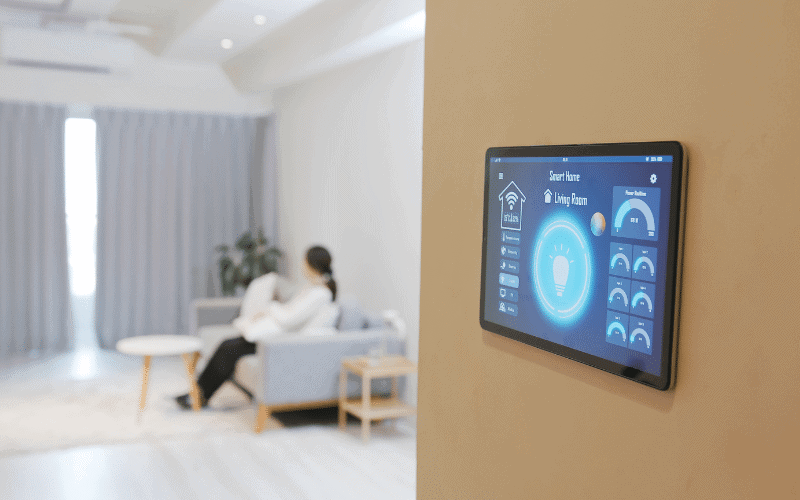 A smart home control panel on a wall, with a cozy living room in the background, features a person enjoying the benefits of a smart thermostat while relaxing on the couch.