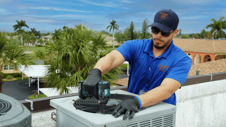 A technician wearing gloves and a cap uses a power drill to work on an outdoor air conditioning unit, surrounded by palm trees.