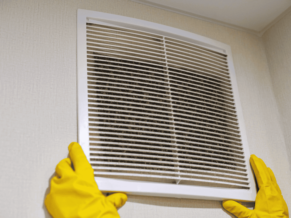 A person with yellow gloves holds a dirty air vent cover on the wall, likely inspecting it for emissions or weird odors affecting the heating system.