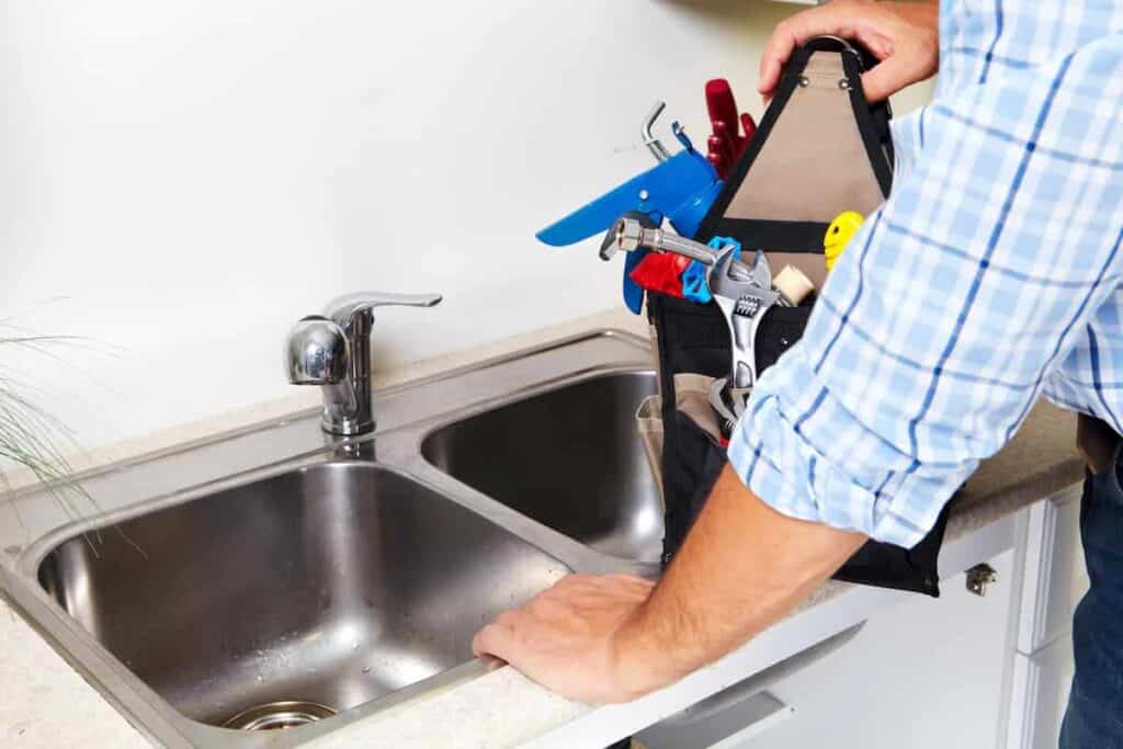 A person holding a toolkit near a kitchen sink is ready to tackle common kitchen clogs, preparing to perform essential plumbing work.