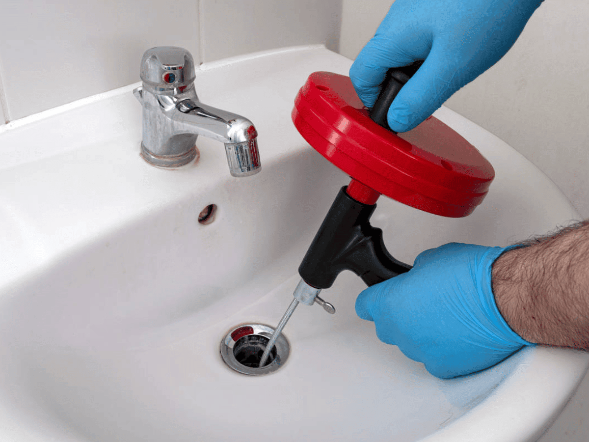Wearing blue gloves, a person uses a red and black drain auger to tackle the stubborn bathroom sink. For those daunting clogged drains, it's wise to call a pro for expert plumbing services.