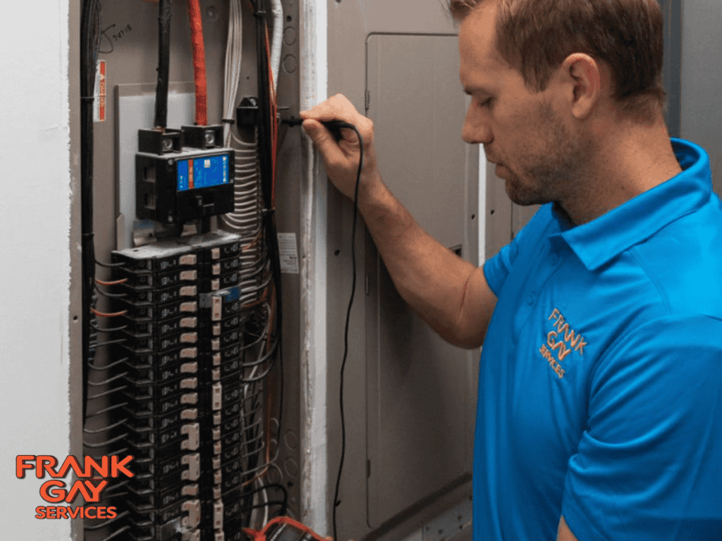 A person in a blue shirt labeled "Frank Gay Services" using a tool to check an electrical panel with many wires.