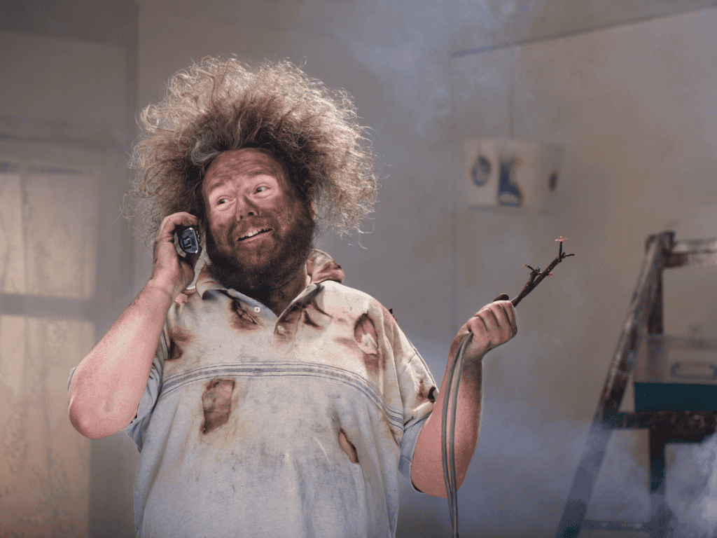 In a smoky room with a ladder in the background, a person with messy hair and a dirty shirt holds a phone and wire, seemingly contemplating an upgrade to the outdated electrical system.