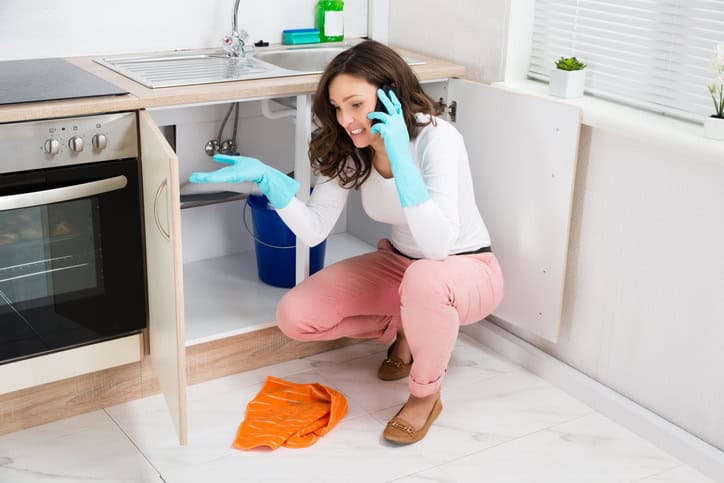 A woman wearing gloves talks on the phone while inspecting under a kitchen sink, highlighting the value of regular plumbing maintenance; a bucket and cleaning cloth are visible by her side.