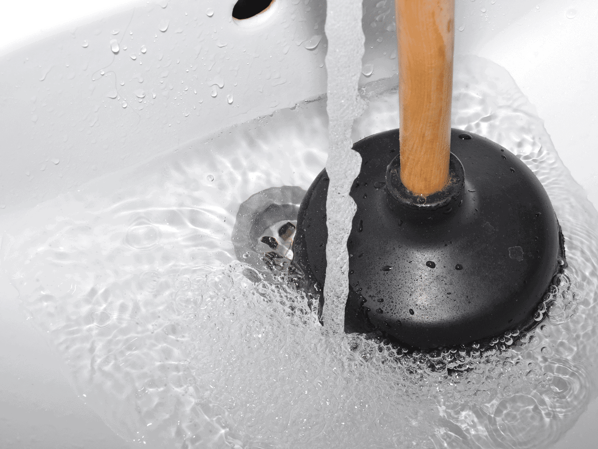 A sink with running water struggles with a clogged drain as a plunger springs into action, showcasing the benefits of regular drain maintenance.