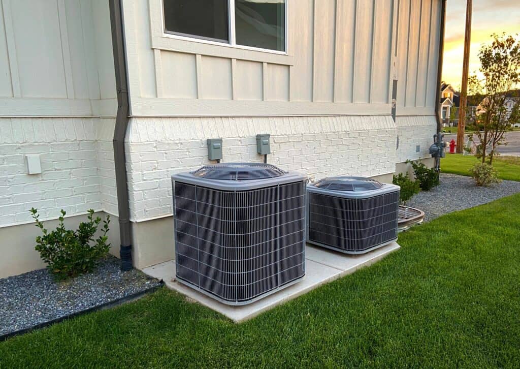 Two outdoor HVAC units are installed on a concrete slab beside a white house, with a grassy lawn in the foreground. As fall approaches, it's the perfect time to schedule maintenance and ensure everything runs smoothly through the cooler months ahead.