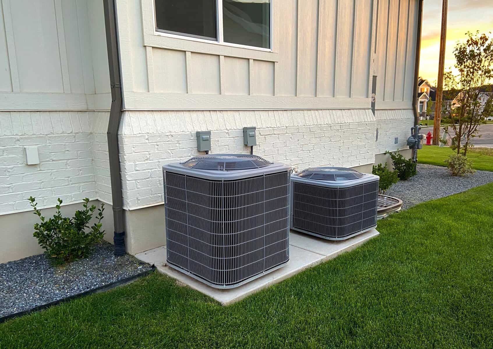 Two outdoor HVAC units are installed on a concrete slab beside a white house, with a grassy lawn in the foreground. As fall approaches, it's the perfect time to schedule maintenance and ensure everything runs smoothly through the cooler months ahead.