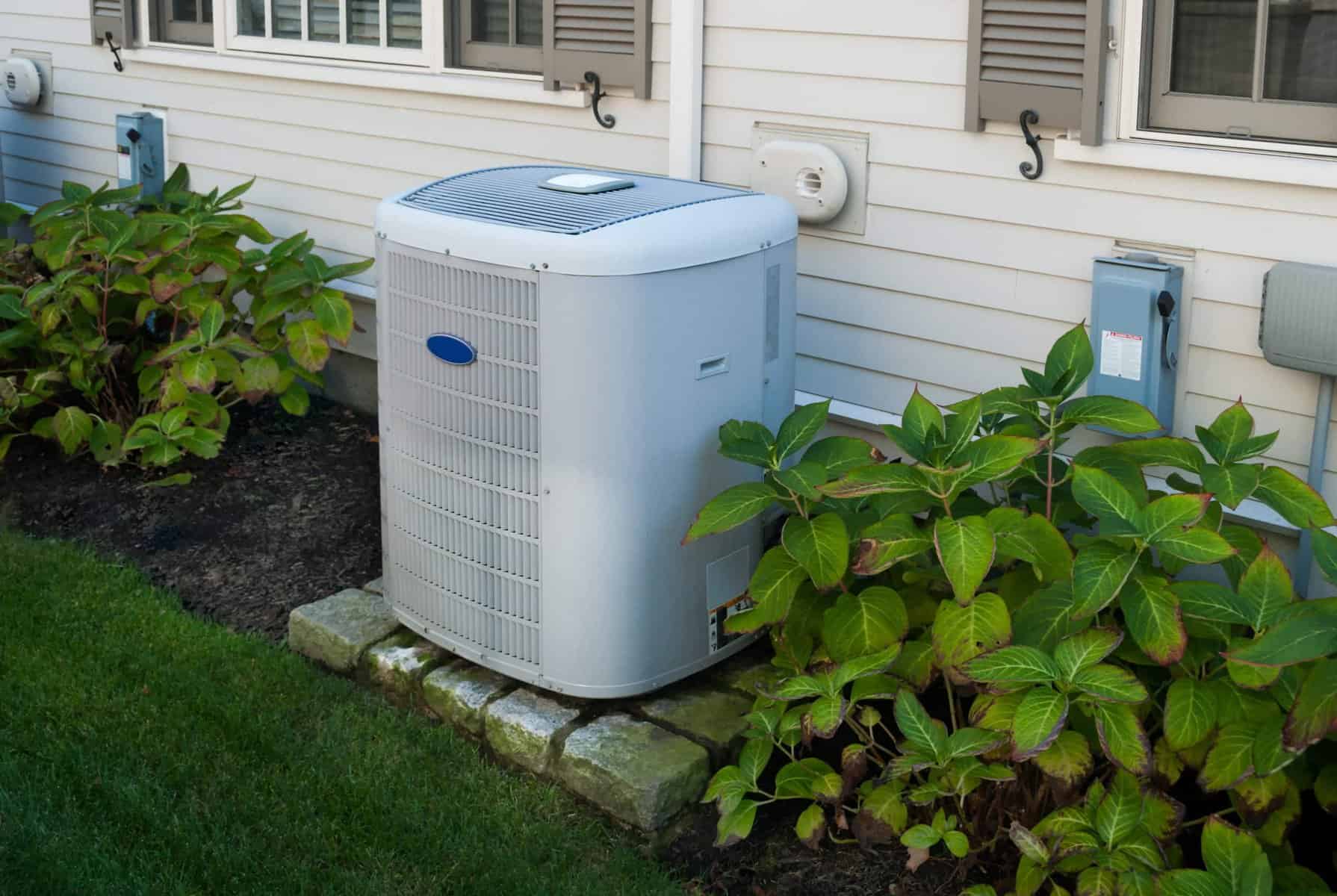 An outdoor air conditioning unit is expertly installed next to a house, surrounded by lush green foliage and grass, highlighting the importance of choosing the right AC to ensure your home system perfectly complements its natural surroundings.