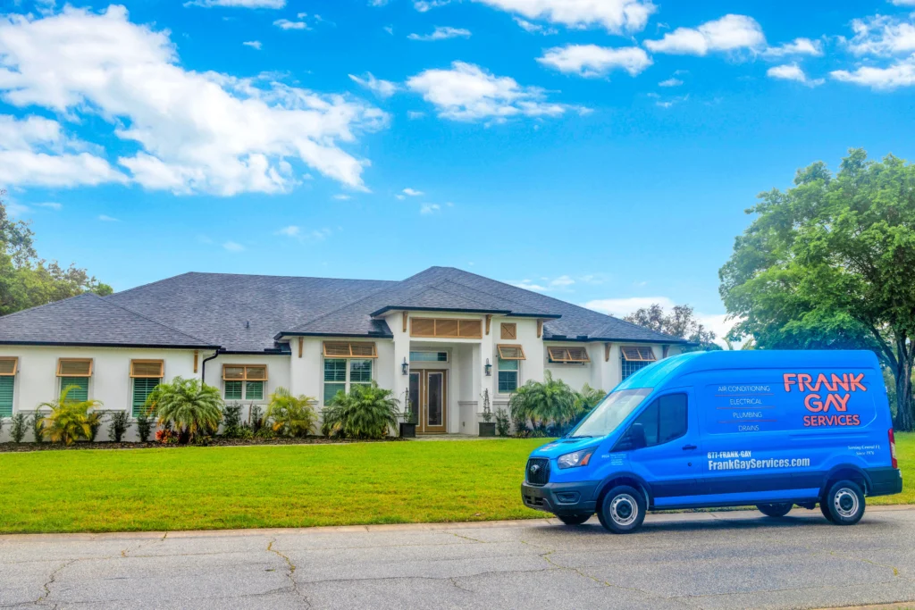 A blue service van labeled "Frank Gay Services" is parked on the street in front of a large, single-story white house with a manicured lawn on a sunny day, reflecting our commitment to quality and community care. Learn more about us by visiting our website or contacting our friendly team.