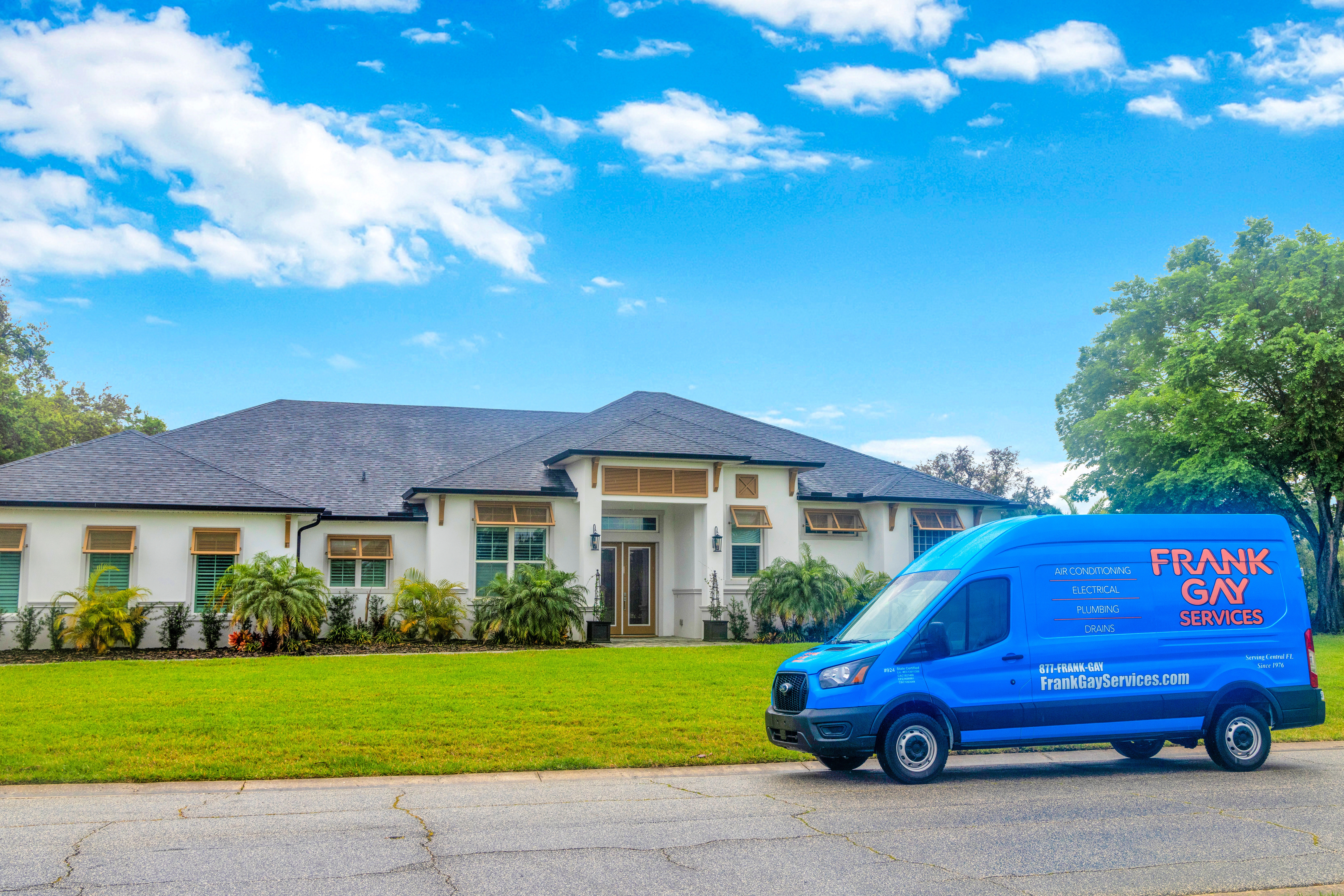 A blue service van labeled "Frank Gay Services" is parked on the street in front of a large, single-story white house with a manicured lawn on a sunny day, reflecting our commitment to quality and community care. Learn more about us by visiting our website or contacting our friendly team.