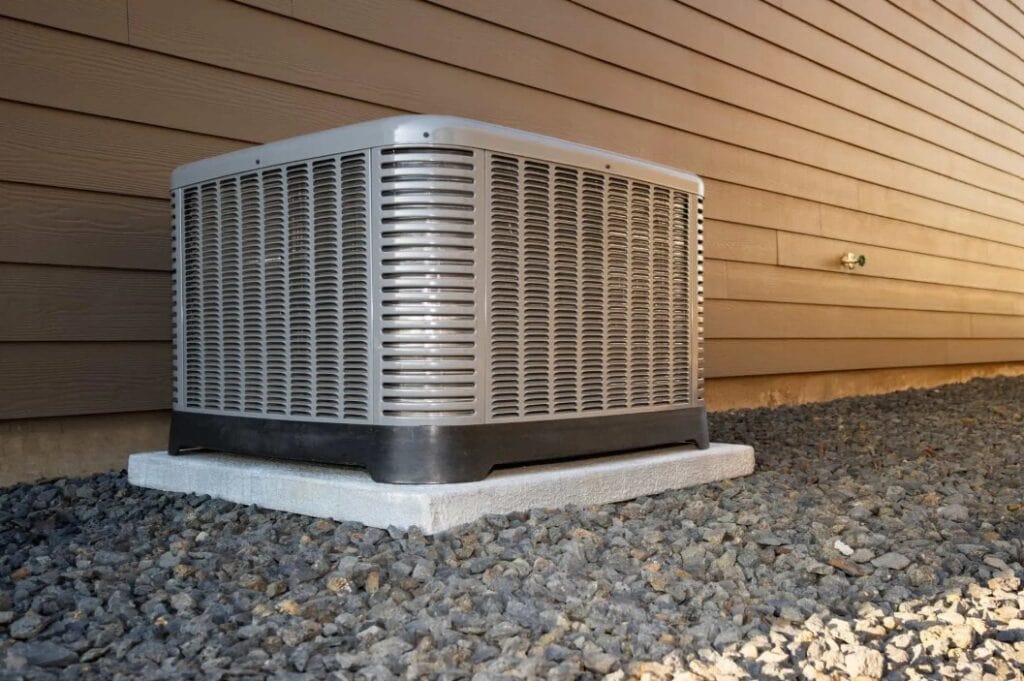 A central HVAC system sits on a concrete pad next to the exterior wall of a house with horizontal siding and a gravel ground surface, aiding in hurricane preparedness by being securely installed.