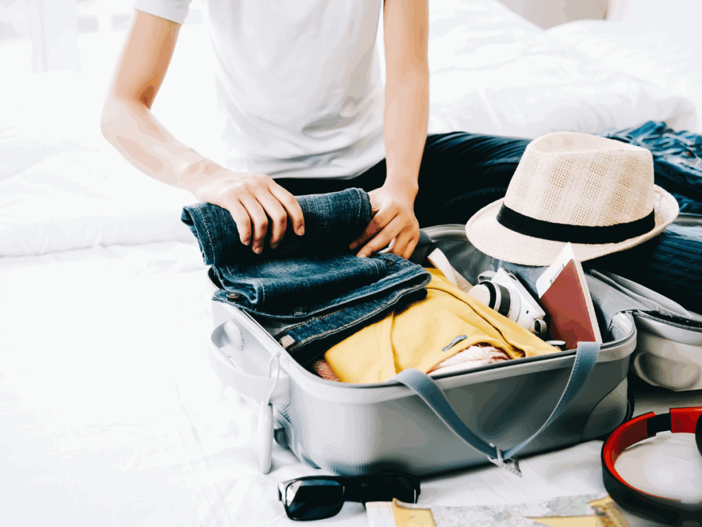 A person is packing clothes into a suitcase on a bed, with a hat, headphones, passport, map, and sunglasses nearby—an essential step in vacation home preparation.