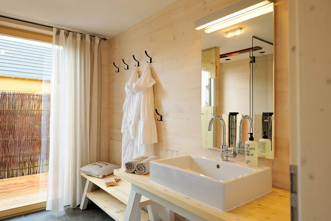 Ein modernes Badezimmer mit quadratischem Waschbecken und großem Spiegel. Auf einer Holzbank liegen gefaltete Handtücher und ein Bademantel. An den Wandhaken hängen Bademäntel. Eine Glastür öffnet sich zu einer Terrasse, und transparente Vorhänge lassen das Tageslicht herein.