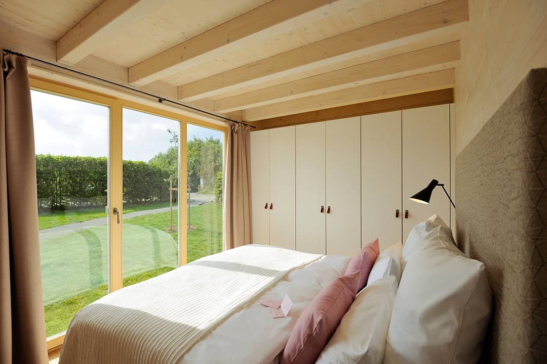 Helles Schlafzimmer mit Holzdecke, großen Glasschiebetüren und Blick auf eine grüne Wiese. Das Bett ist mit weißer und rosa Bettwäsche ausgestattet, und rechts befindet sich eine Reihe weißer Schränke mit braunen Griffen. Über dem Bett hängt eine schwarze Leselampe.
