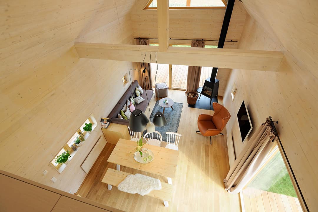 Luftaufnahme eines gemütlichen, modernen Wohnraums mit Holzinterieur. Er verfügt über einen Esstisch mit Stühlen und einer Bank, einen Plüschsessel, ein Sofa, einen Holzofen und dekorative Pflanzen. Große Fenster sorgen für natürliches Sonnenlicht.