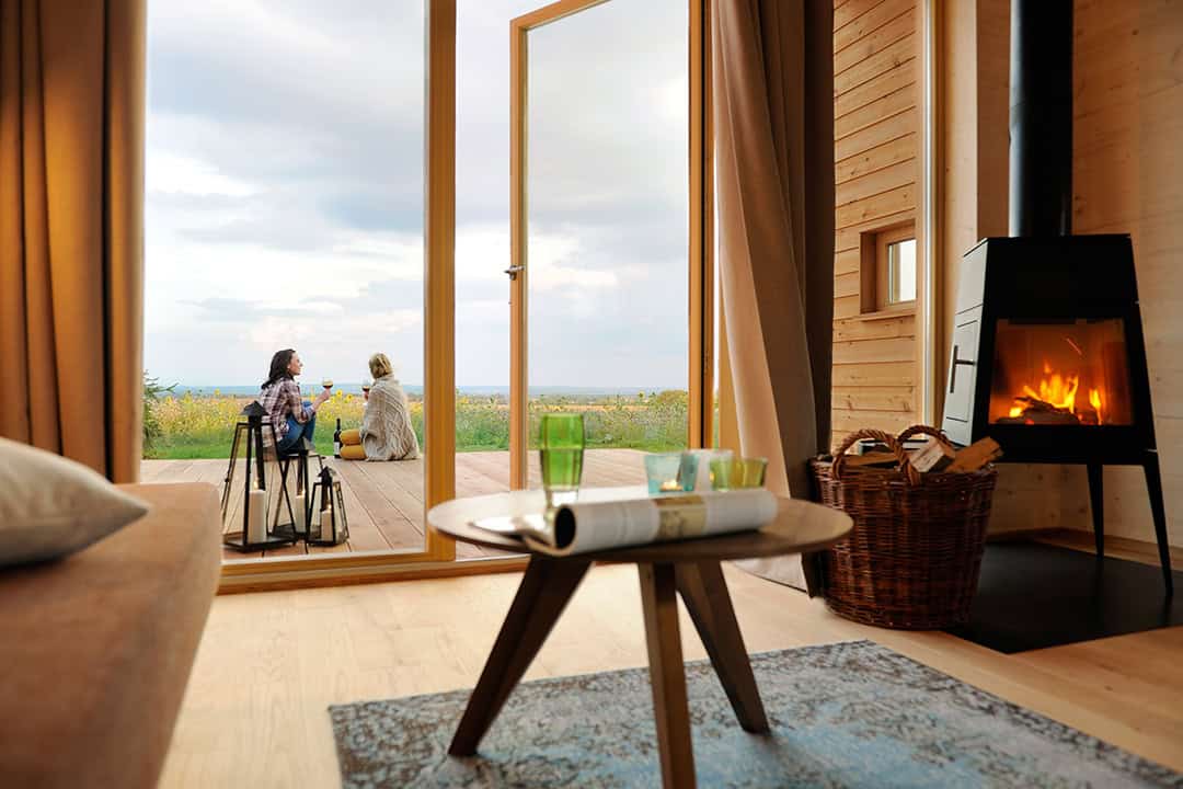 Zwei Personen sitzen auf einer Holzterrasse und genießen durch eine offene Tür den Blick auf ein Feld unter bewölktem Himmel. Drinnen erwartet Sie ein gemütlicher Raum mit einem brennenden Kamin, einem Couchtisch mit Gläsern und einer Zeitung sowie großen Fenstern.