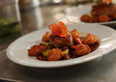 Ein Gourmetgericht auf einem weißen Teller mit einem Stück Fleisch, garniert mit essbaren orangefarbenen Blüten und Kräutern, umgeben von kunstvoll aufgespritztem Kartoffelpüree und einer reichhaltigen Sauce. Der Hintergrund ist sanft verschwommen und hebt das Gericht hervor.
