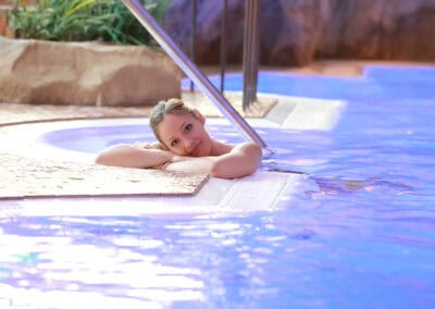 Eine Person mit hellem Haar entspannt sich am Rand eines Swimmingpools und lehnt mit verschränkten Armen am Beckenrand. Das Wasser spiegelt einen violett-blauen Farbton. Der Poolbereich strahlt mit Steindekorationen und Pflanzen im Hintergrund eine ruhige Atmosphäre aus.