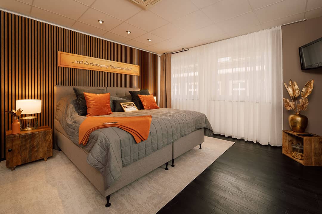 Modernes Schlafzimmer mit großem Bett in Grau und Orange. Eine Holzwand mit Leuchtreklame ziert ein Nachttisch mit Lampe und Pflanze. Transparente Vorhänge verdecken ein breites Fenster. Dekorative goldene Stehlampen.
